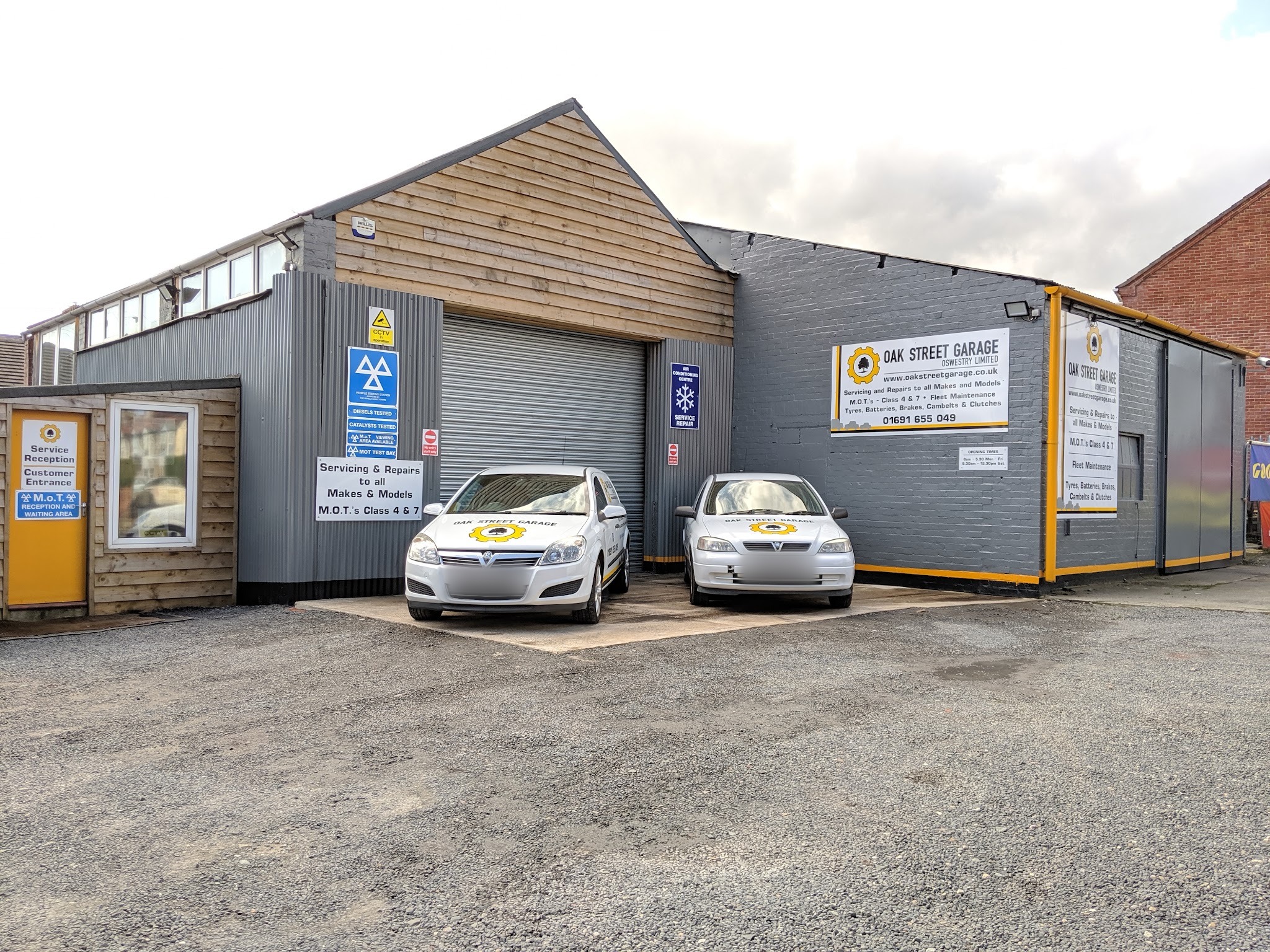 The Oak Street Garage workshop: grey cladding, a roller door, MOT and air conditioning signs, the reception cabin and two cars in Oak Street Garage livery